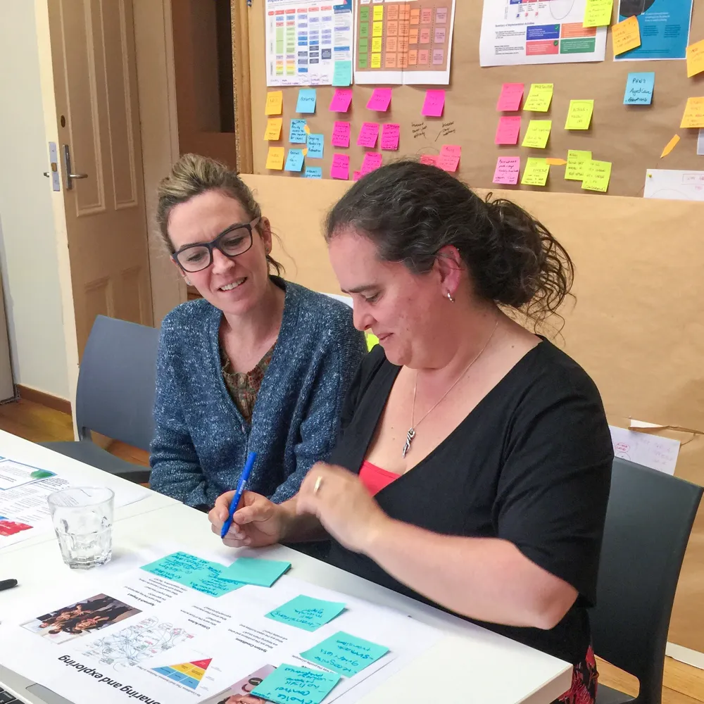 Two women sit at a table covered with papers and sticky notes, collaborating. A bulletin board with colourful notes is visible in the background.