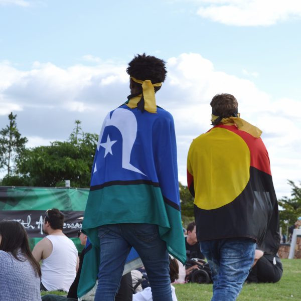 Two people walking through a crowd with the Aboriginal flag and Torres Strait Islander flag draped over their shoulders.