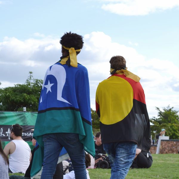 Two people walking across a lawn with an Aboriginal flag and Torres Strait Islander flag draped over their shoulders.