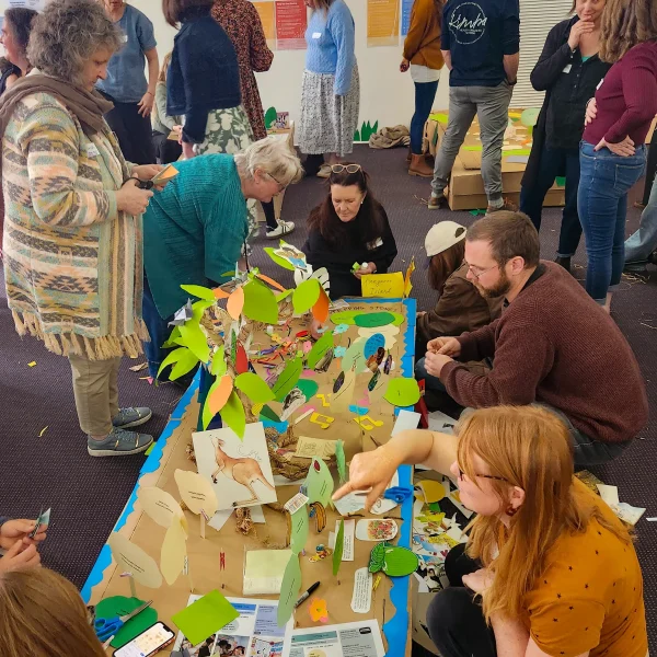 A group of adults gathered around a large table on the floor working together on a colourful craft workshop piece: cut-out paper leaves and shapes, a model tree, kangaroo illustration, and other materials spread across the table as participants add and assemble elements.