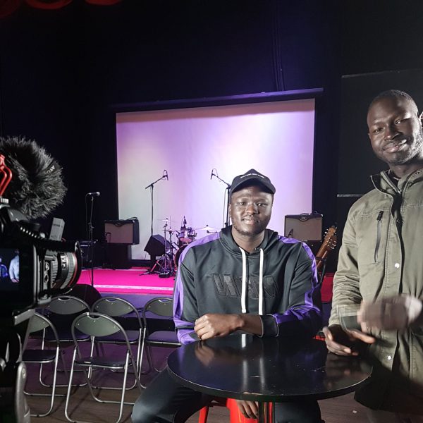Two people with South Sudanese backgrounds sitting in front of a stage with a video camera.